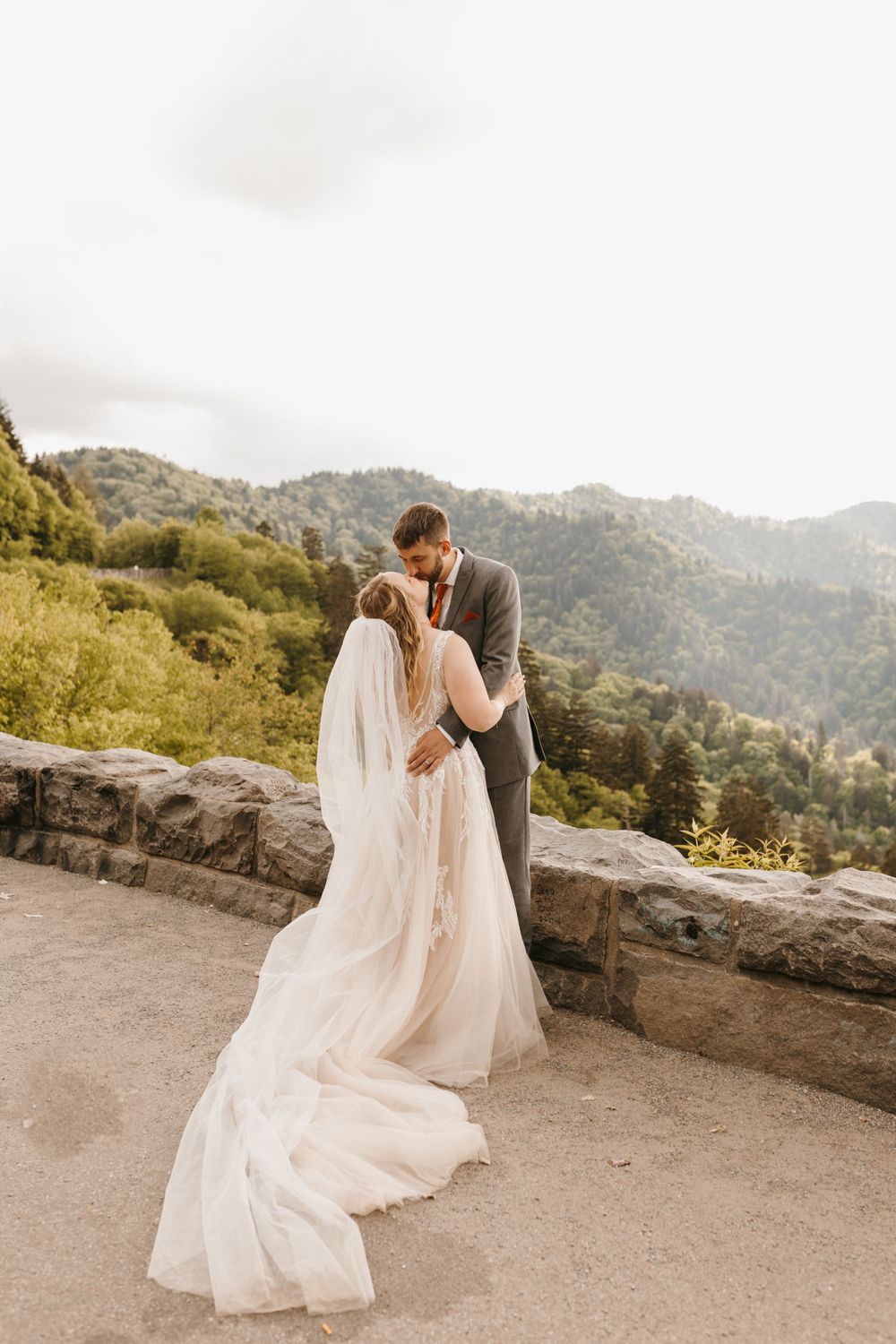 Bride and groom kiss tenderly with flowing veil and train at mountain overlook elopement.