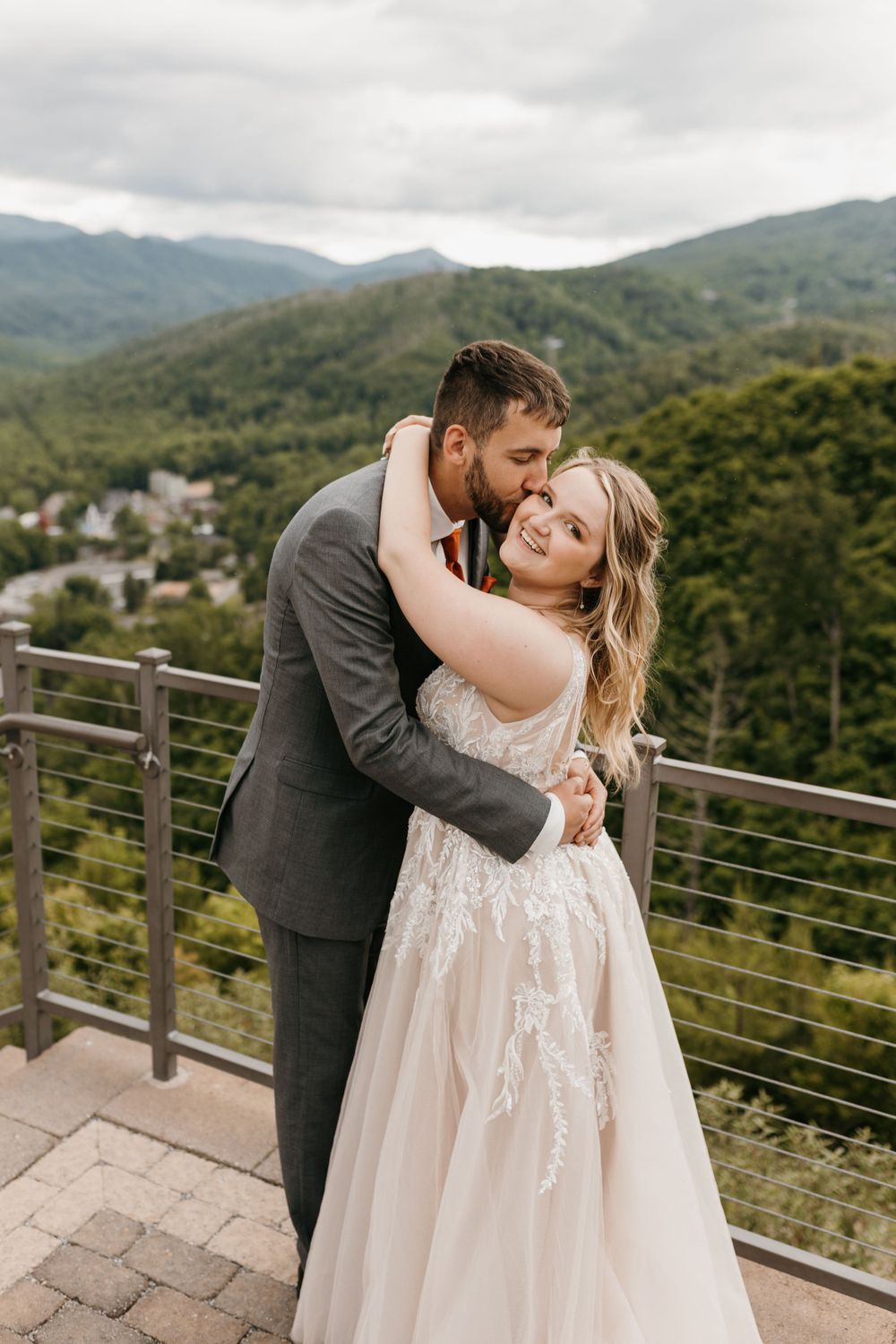 Groom embraces bride from behind with a kiss as she smiles, framed by misty mountain scenery.