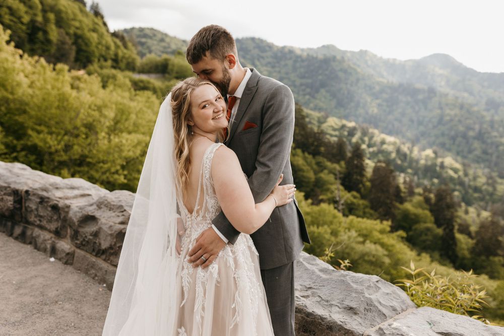 Groom embraces bride in lace veil gown as she smiles at mountain overlook wedding.