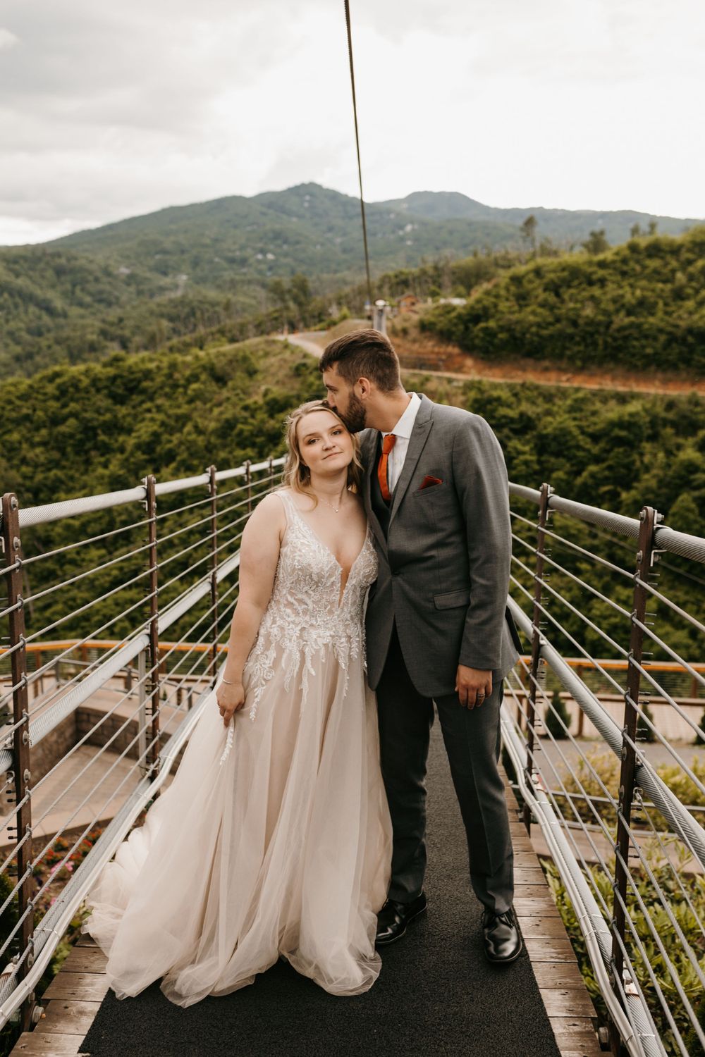 Groom kissing bride on cheek on a mountain suspension bridge with scenic backdrop.