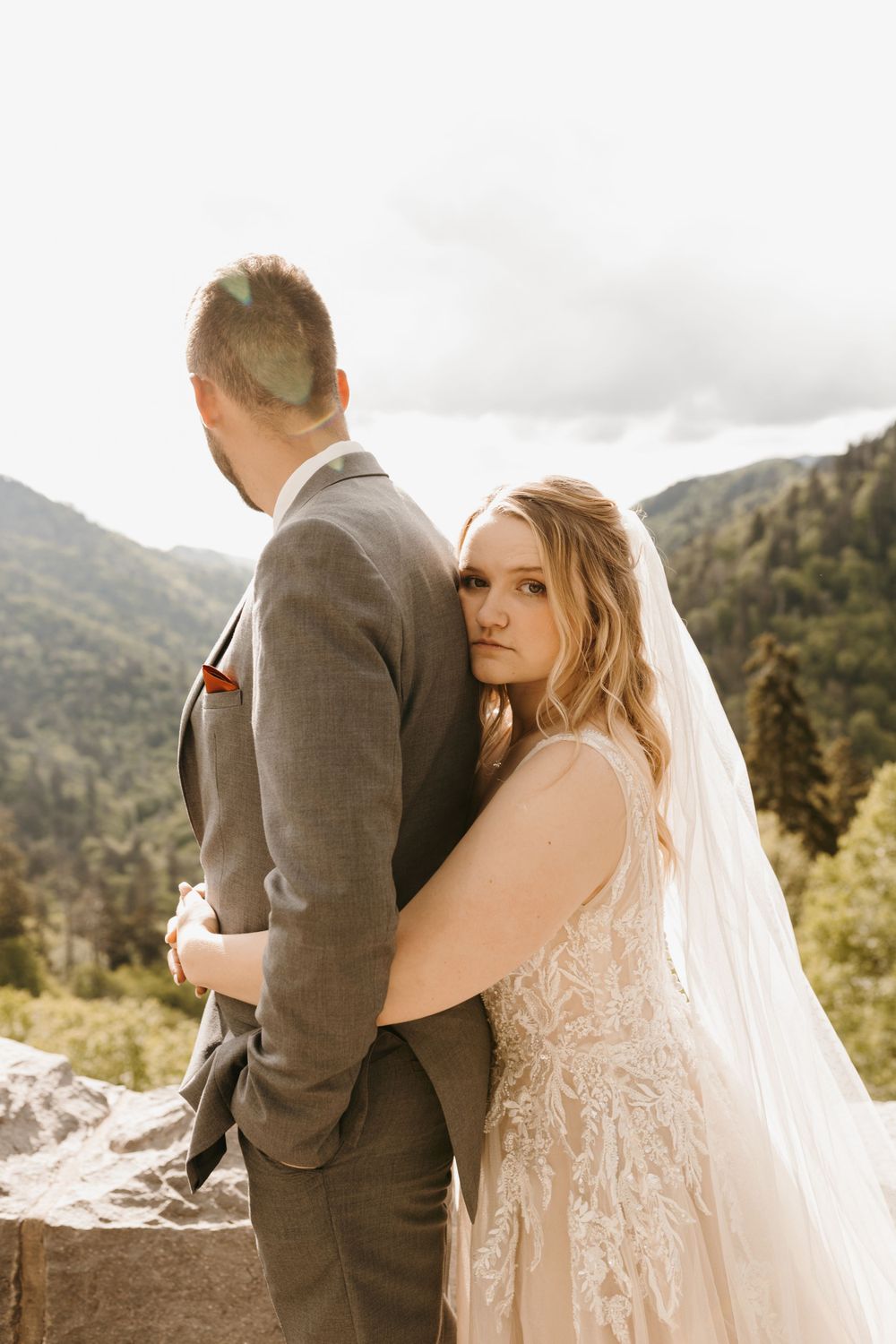 Bride in lace gown leans against groom in gray suit at mountain overlook wedding.