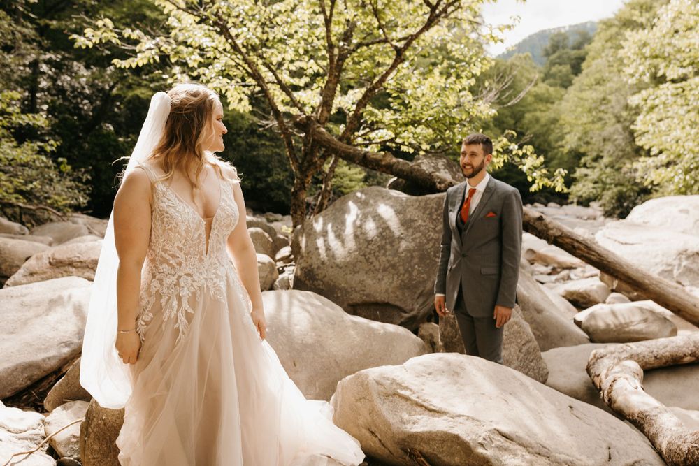 Bride in lace gown and groom in gray suit share a first look moment on a rocky riverbank.