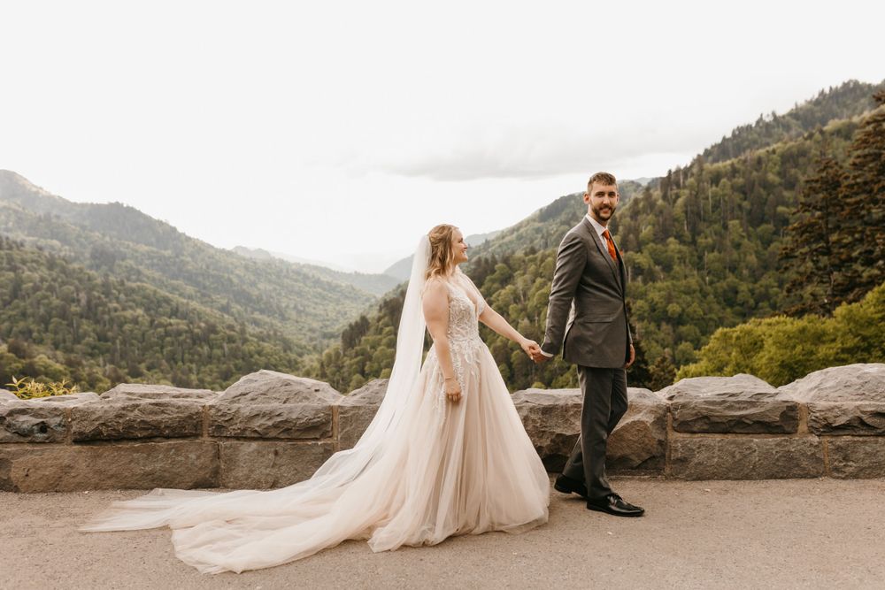 Smiling groom leads bride by hand along stone wall with mountain scenery.