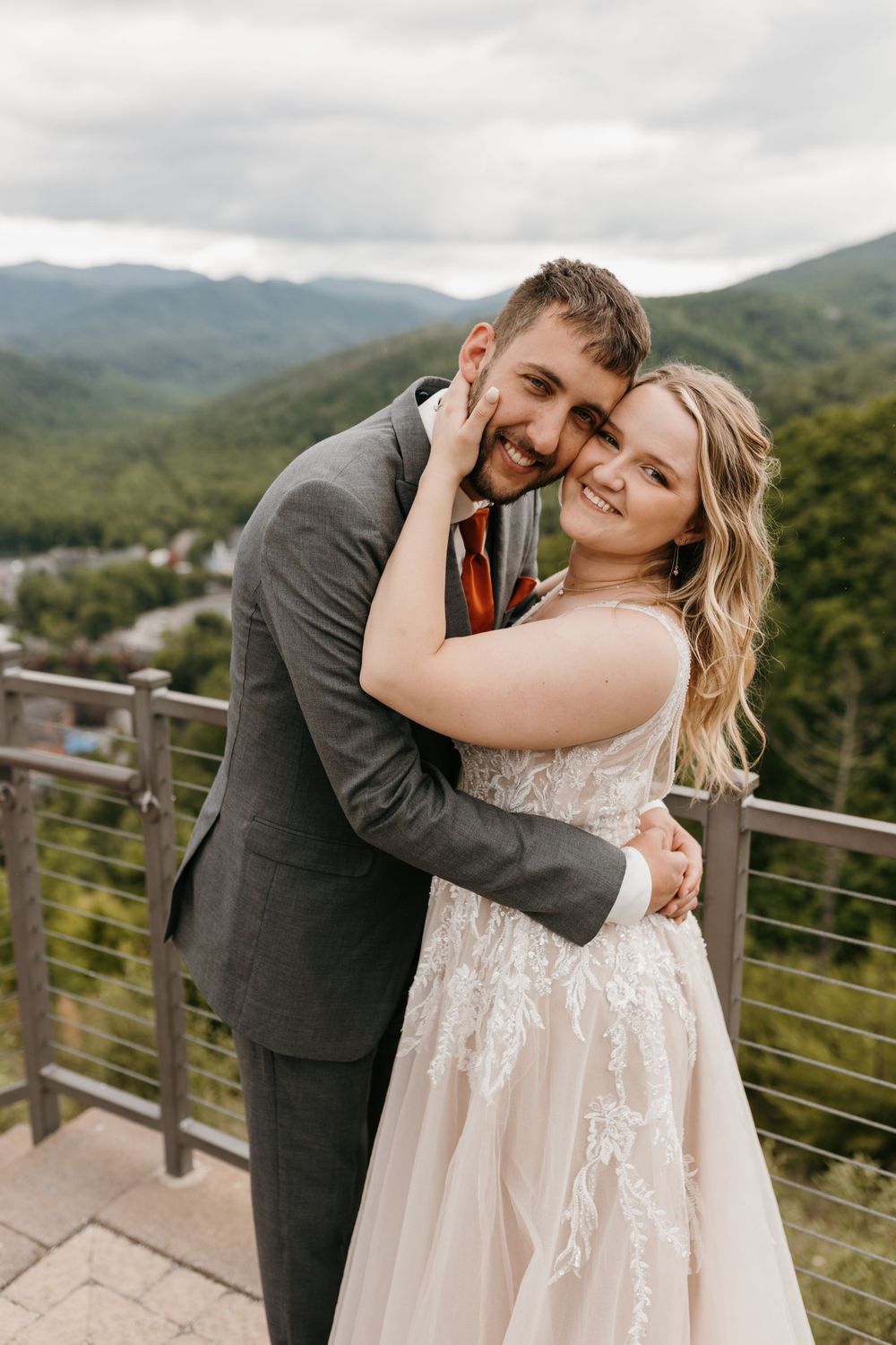 Smiling bride and groom share a tender embrace on an outdoor terrace overlooking a mountain town.