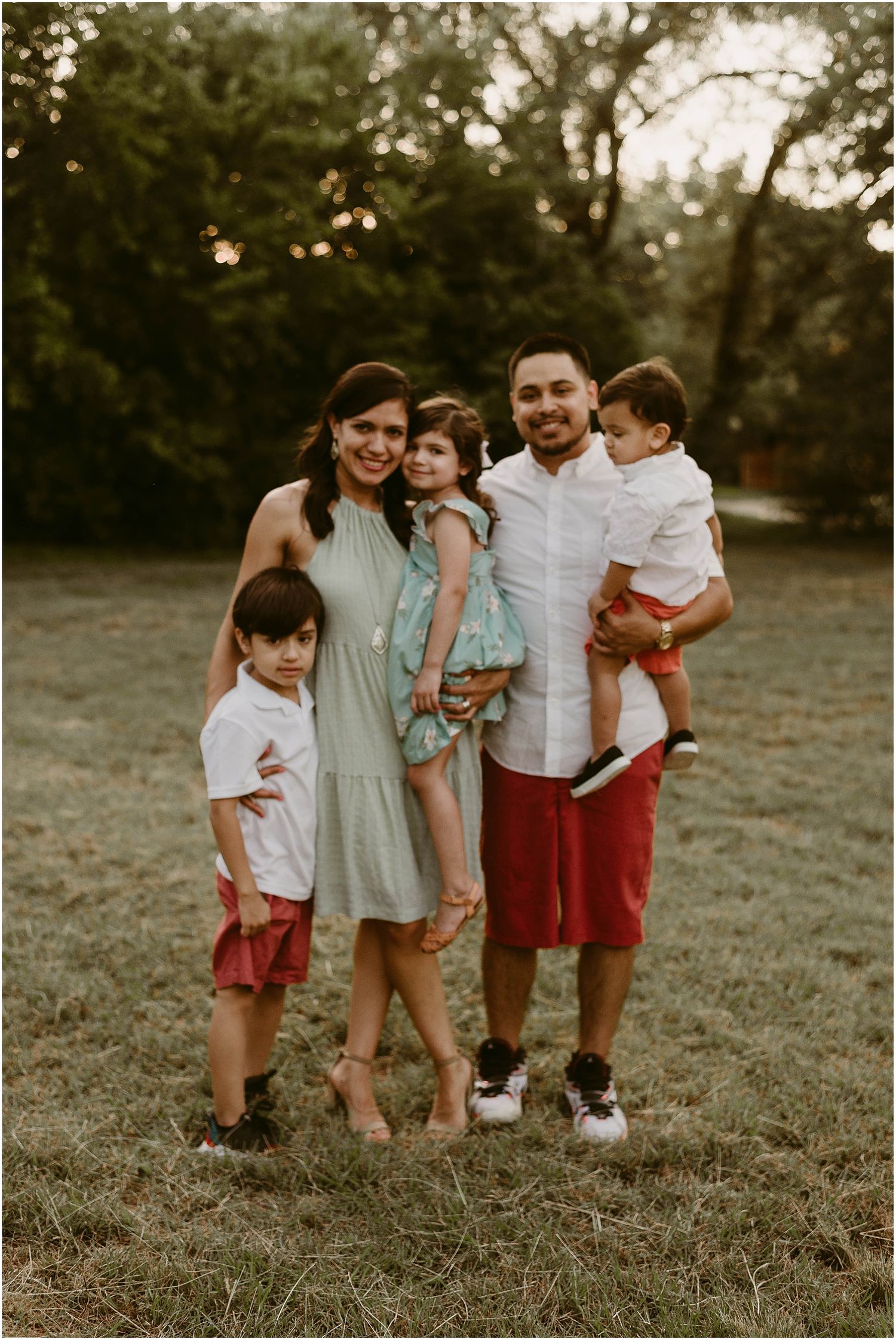 A family poses together outdoors at sunset wearing coordinated casual outfits in white and red tones.