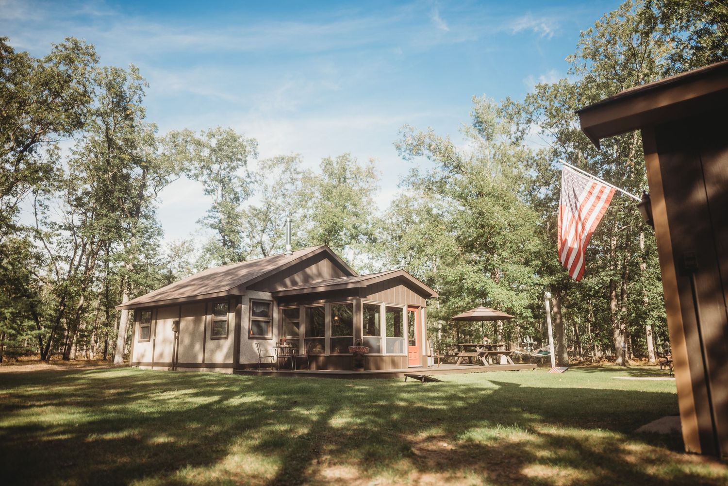 Rural ranch-style house with American flag surrounded by trees under blue sky.