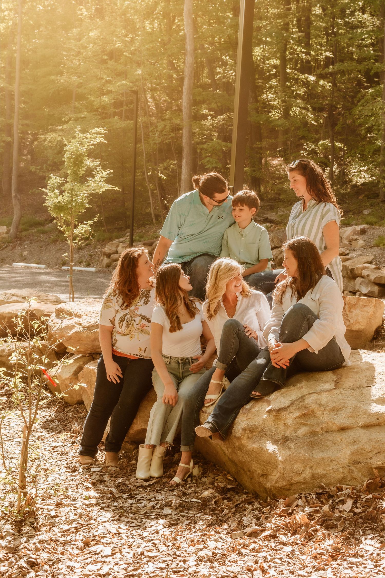A group relaxes together on large rocks in a sunlit wooded area during an outdoor photoshoot.