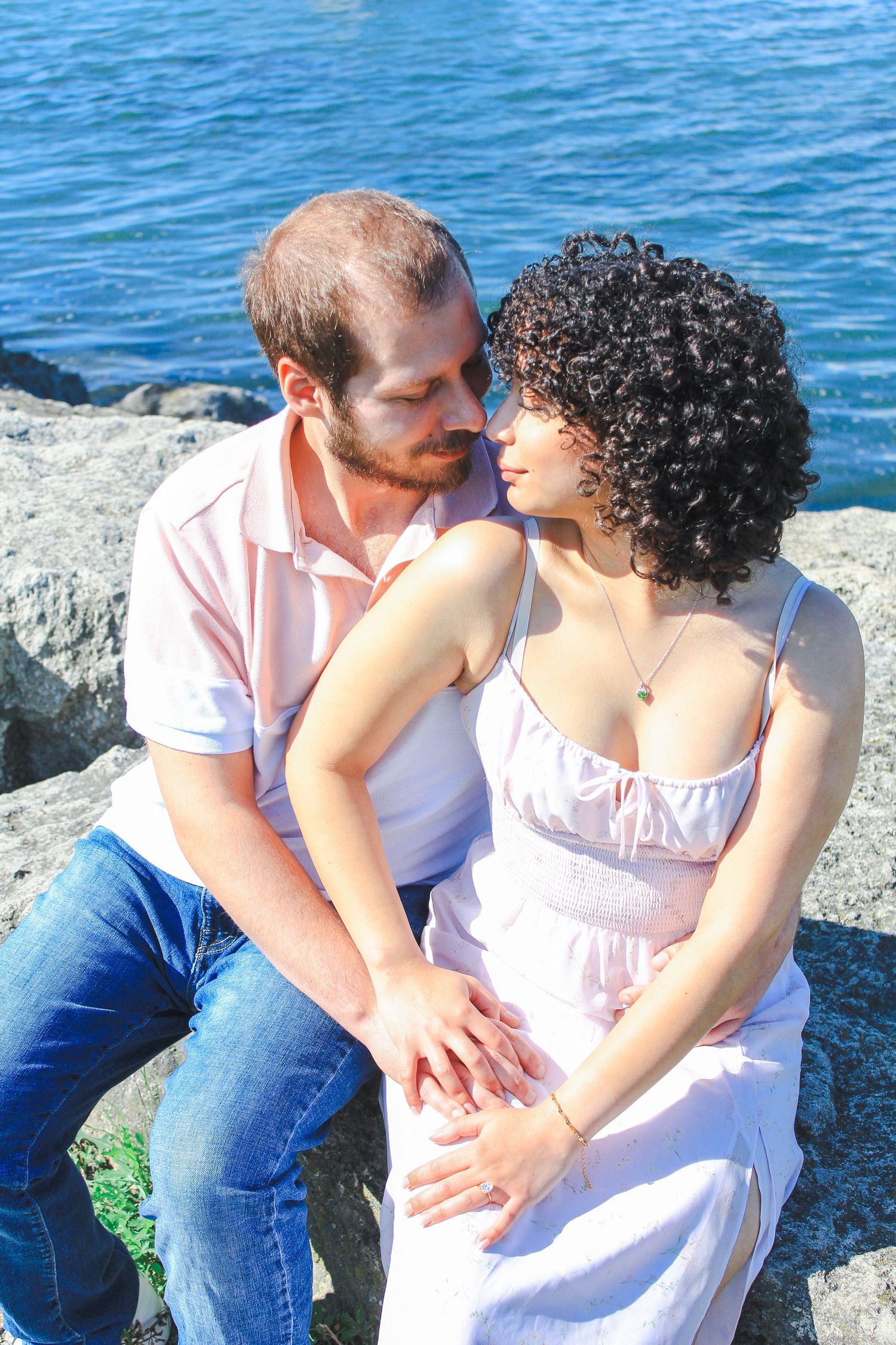 A romantic couple sits together on coastal rocks with the blue ocean in the background.