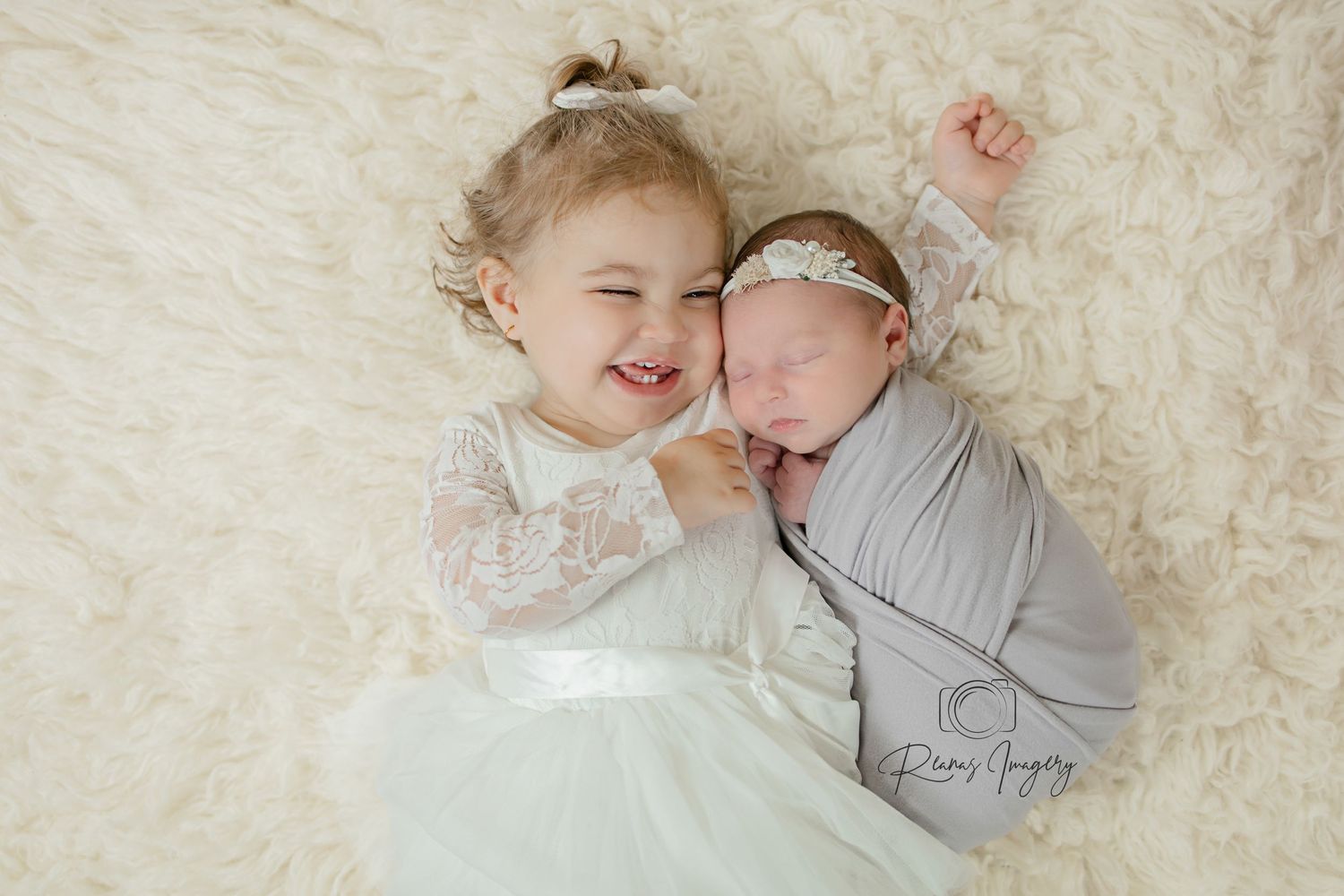 Smiling toddler girl in white lace dress cuddling with swaddled newborn sibling on fur rug.