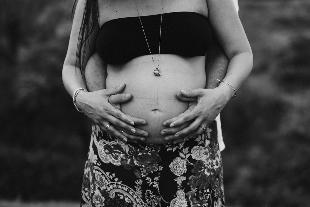 Black and white photo of a pregnant belly with a delicate necklace against patterned fabric.