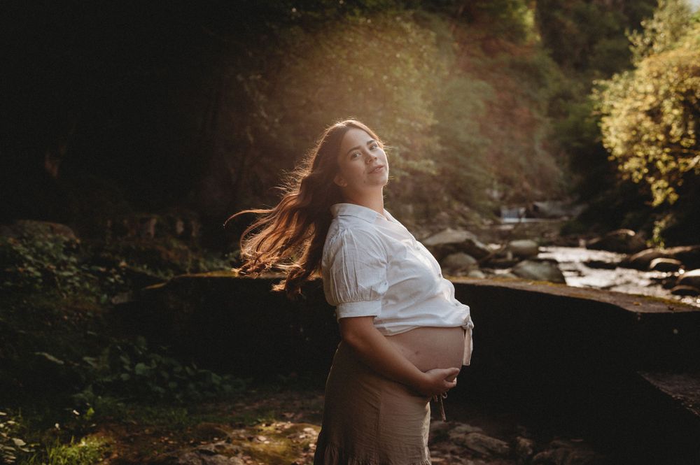 Maternity portrait in white dress against rocky stream backdrop with sunlight.