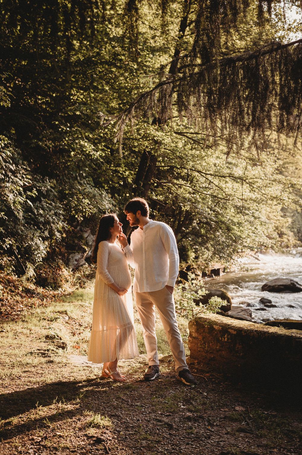 A romantic couple in white outfits sharing an intimate moment by a stream in a sunlit forest setting.