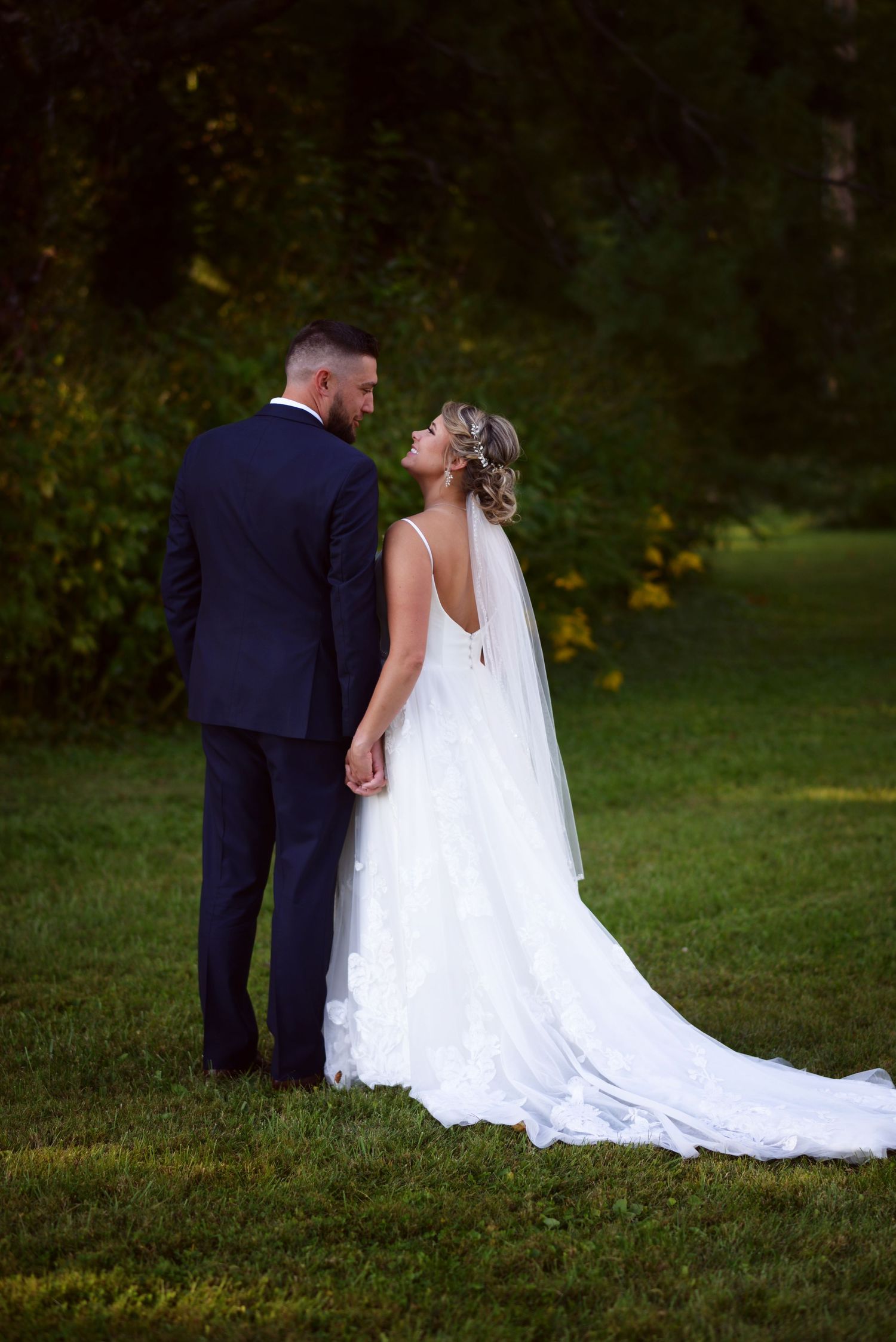 A bride in a flowing white gown and groom in navy suit pose together on grassy lawn at sunset.