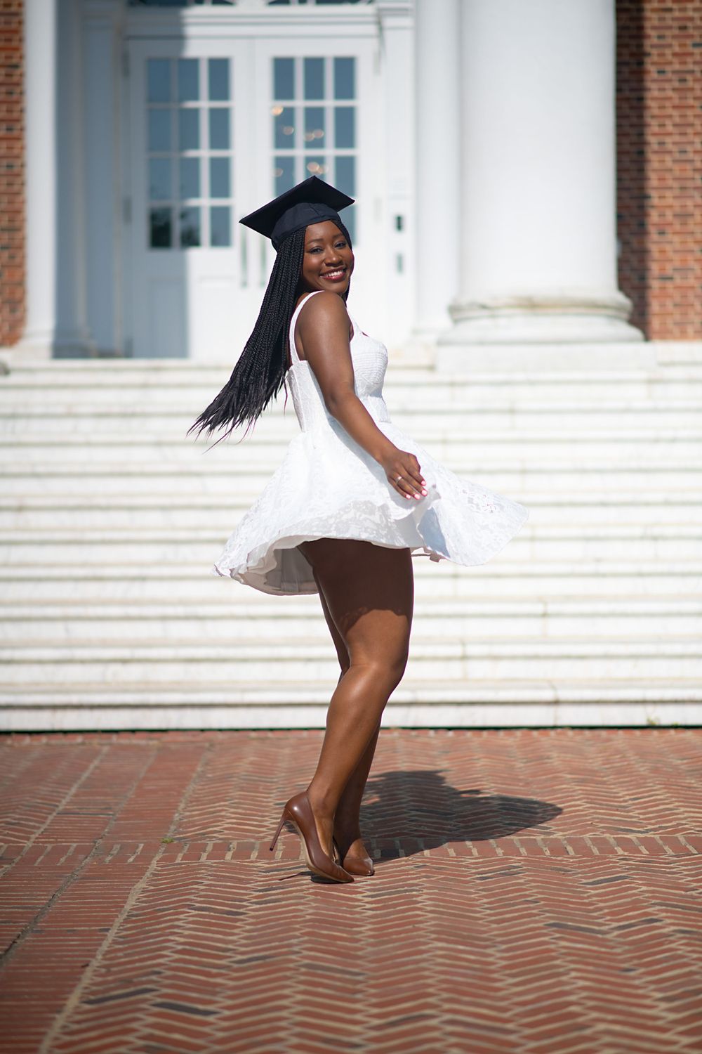 A graduate celebrates on brick steps in front of white columns wearing a cap and gown.