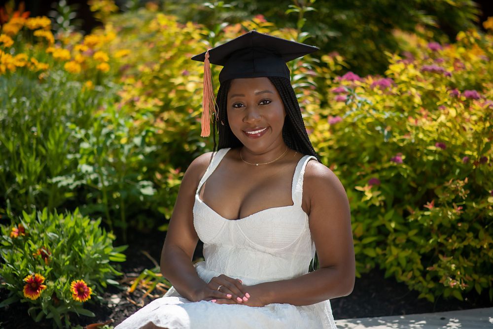 Graduate posing in white dress among yellow flowers and garden foliage.