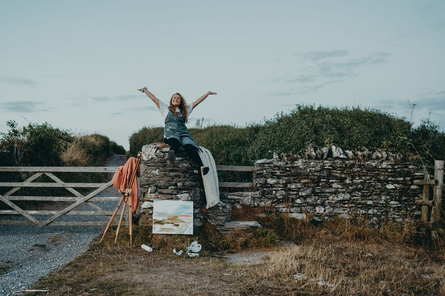 Person sitting on stone wall with art supplies and camera during sunset.
