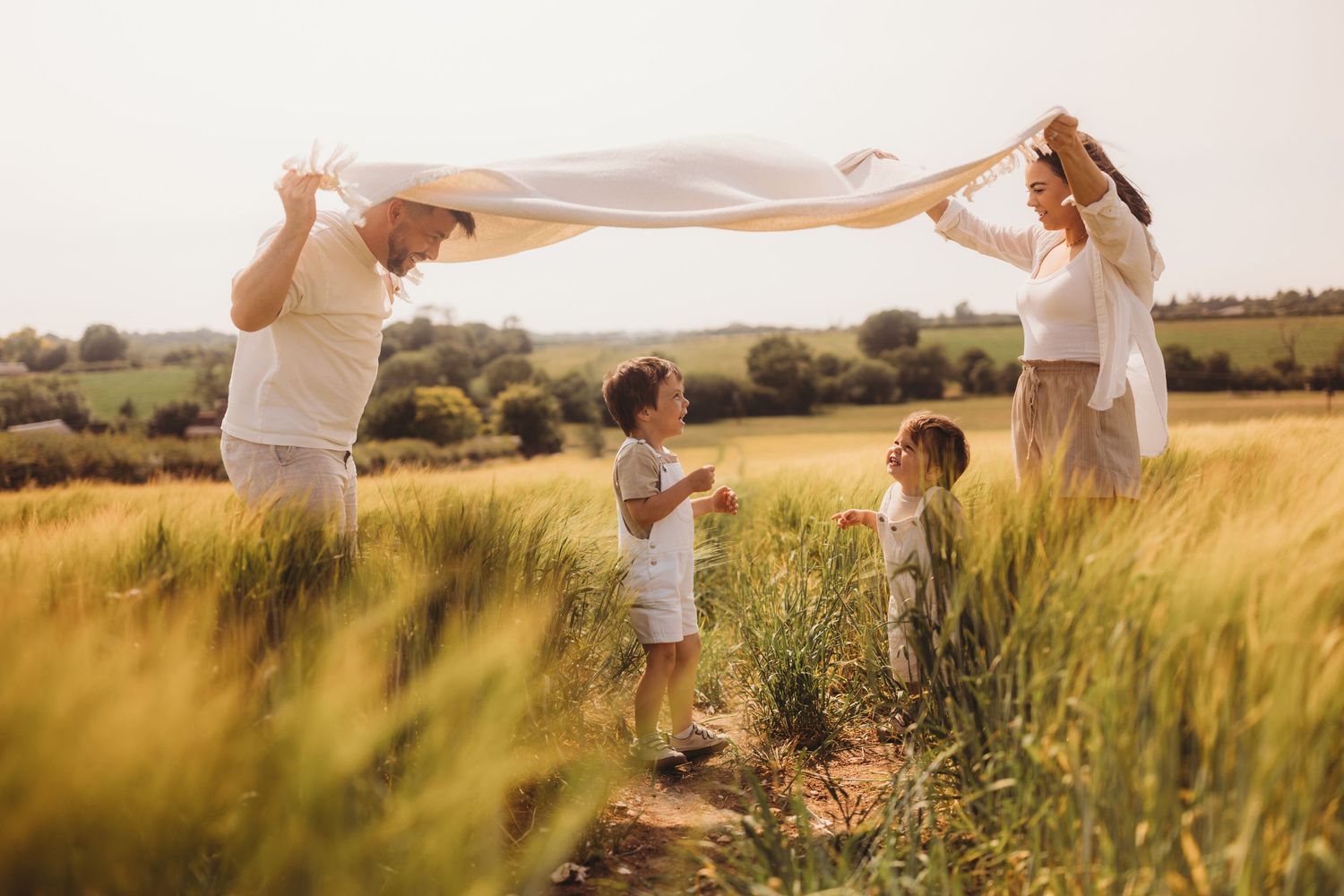 Family playing with white fabric in golden summer field at sunset as wind blows through tall grass.