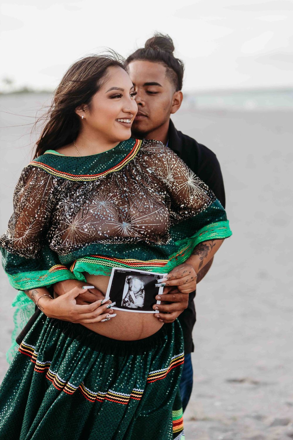 Couple holding ultrasound photo on beach in traditional green and sequined dress.