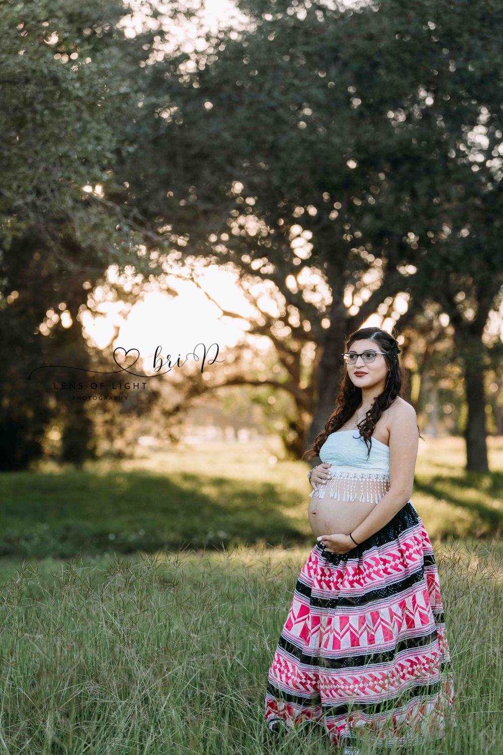 Expectant mother in pink patterned skirt poses in park during sunset maternity session.