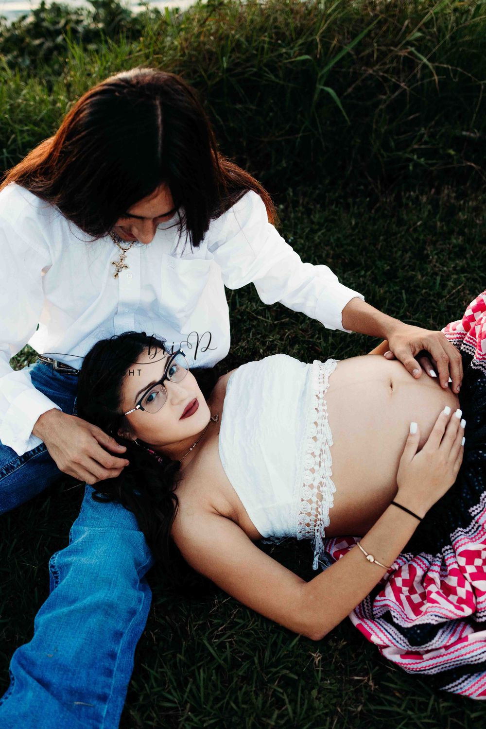 Two people sharing tender moment during outdoor maternity photoshoot on grassy ground.