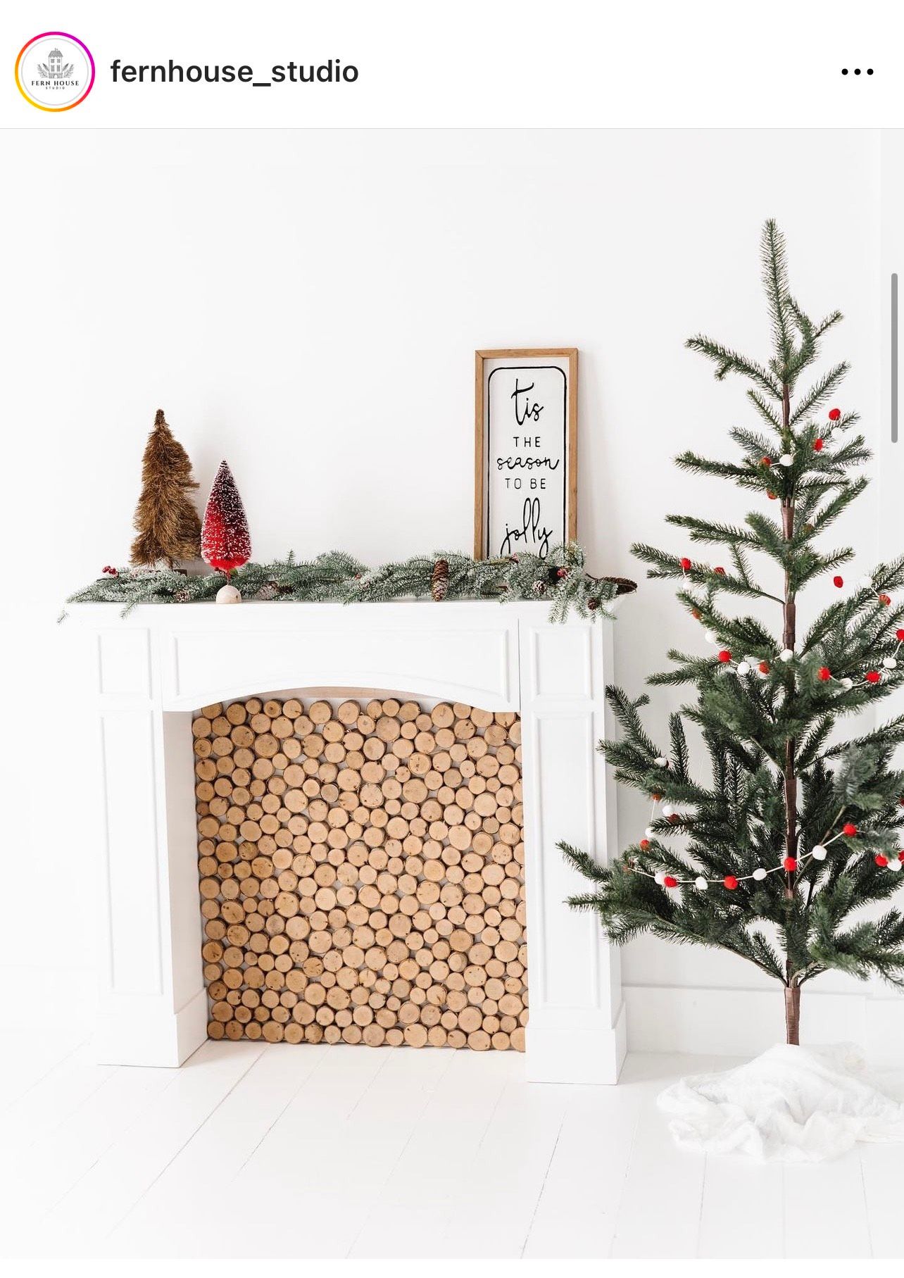 Minimalist Christmas decor with a white fireplace, stacked logs, and sparse decorated tree against white walls.