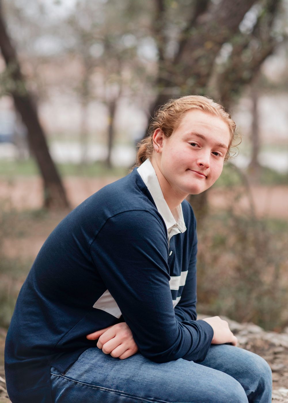 A person wearing a navy sweater and jeans sits outdoors on a rock with a gentle smile on their face.