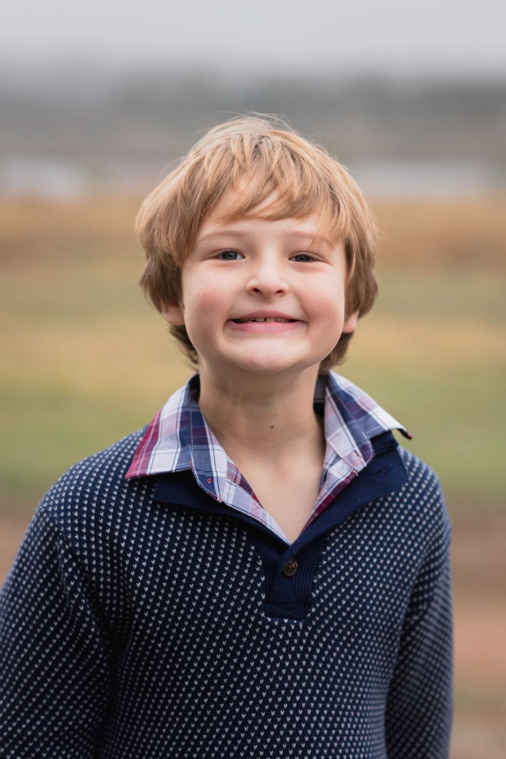 Young child wearing polka dot navy sweater and plaid collar smiles outdoors on foggy day.