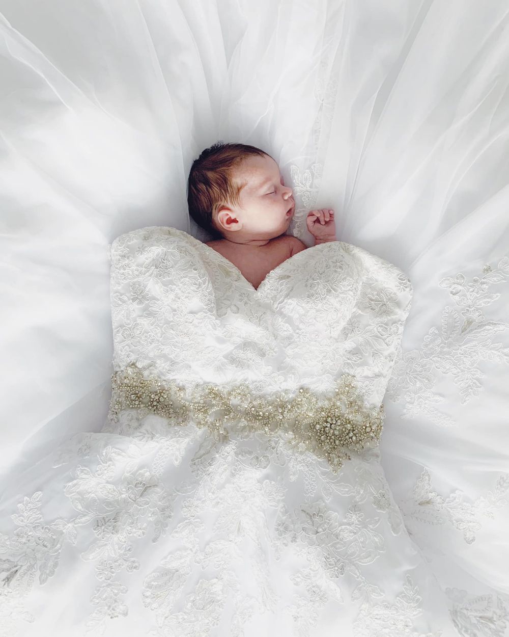 Sleeping newborn nestled in white wedding dress with intricate lace and beaded details.