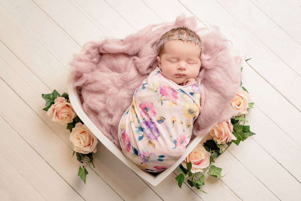 Sleeping newborn wrapped in floral swaddle lying in heart shaped basket surrounded by pink roses.