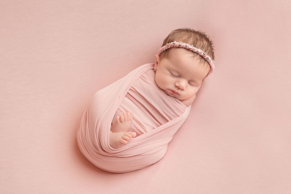Peaceful newborn swaddled in soft pink fabric against pink background with delicate headband.