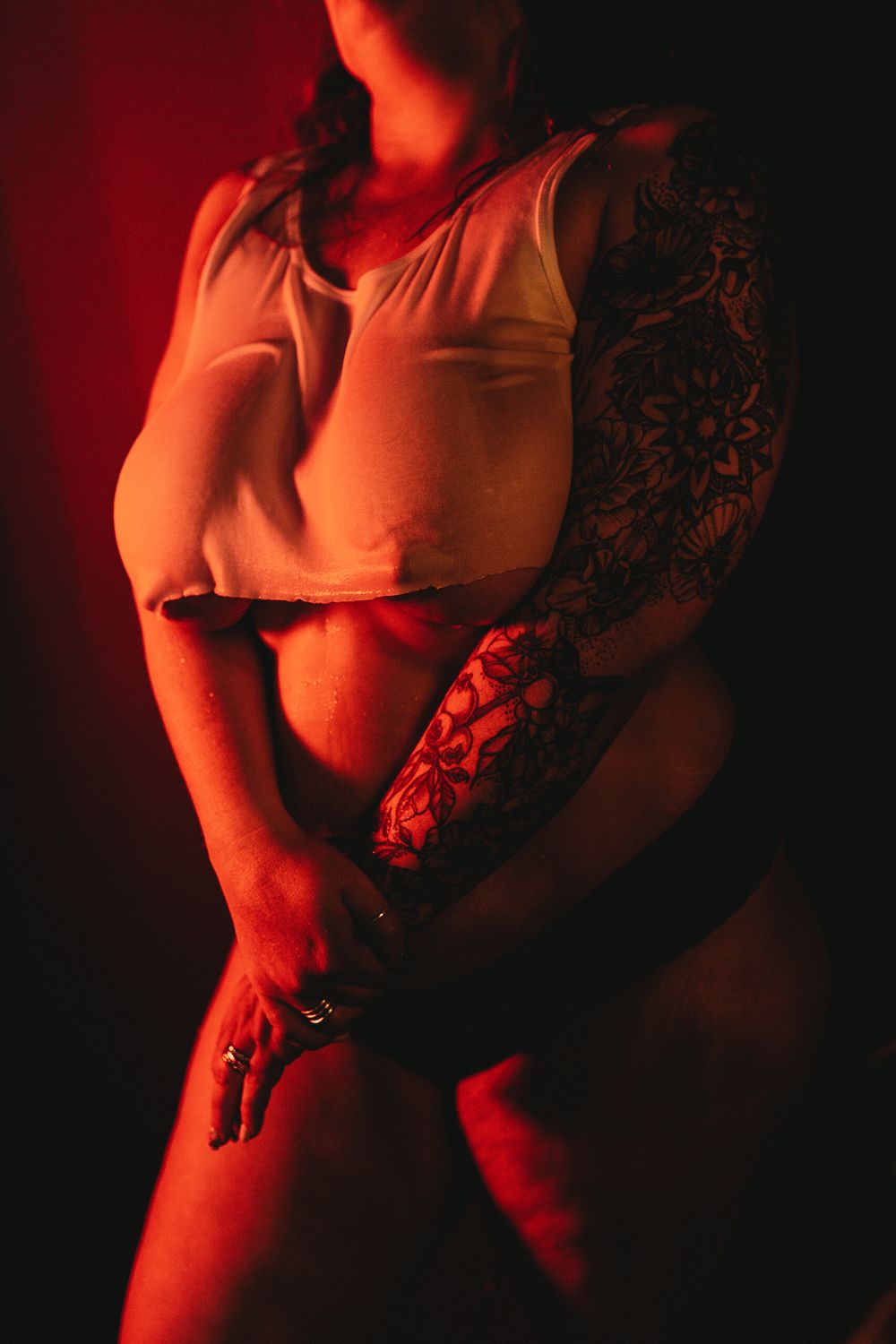 Figure in white tank top with sleeve tattoo poses against red dramatic lighting.