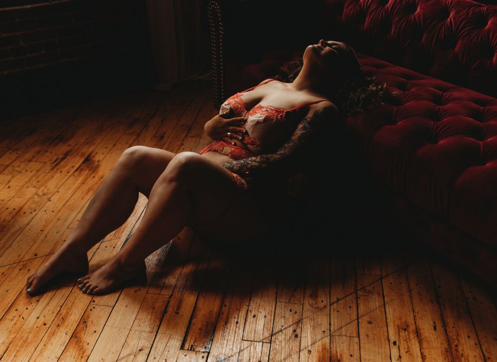 A figure reclines on a plush red velvet couch against wooden flooring in an intimate, dimly lit setting.