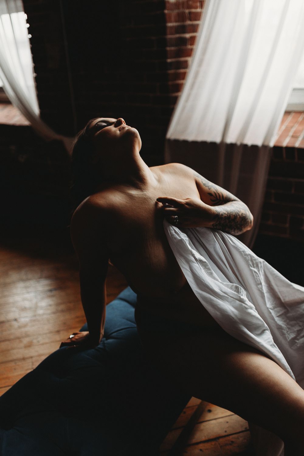 Silhouetted figure in white fabric reclining dramatically against dark wood floor with brick wall and sheer curtains in backdrop.