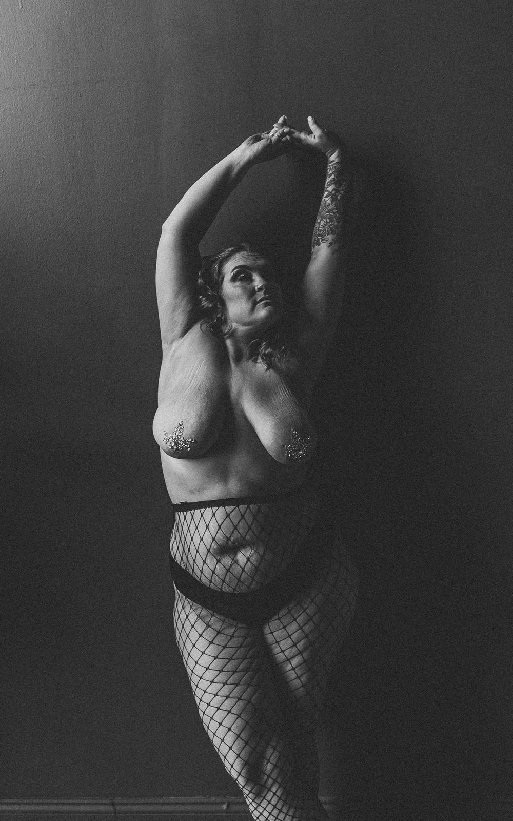 A black and white artistic boudoir photo of a plus size figure posing in fishnet stockings.