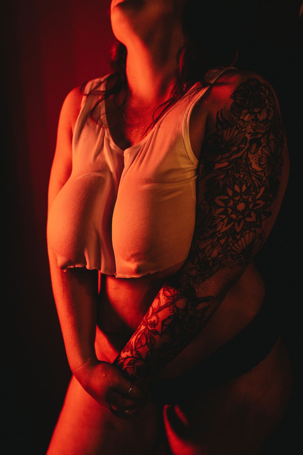 Moody boudoir photo featuring intricate arm tattoo and white tank top in red lighting.
