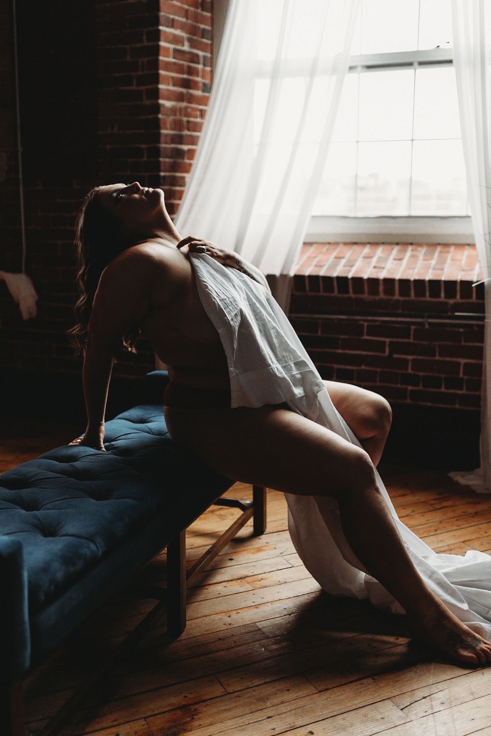 Silhouetted figure lounges near bright window in moody bedroom with exposed brick wall and hardwood floors.