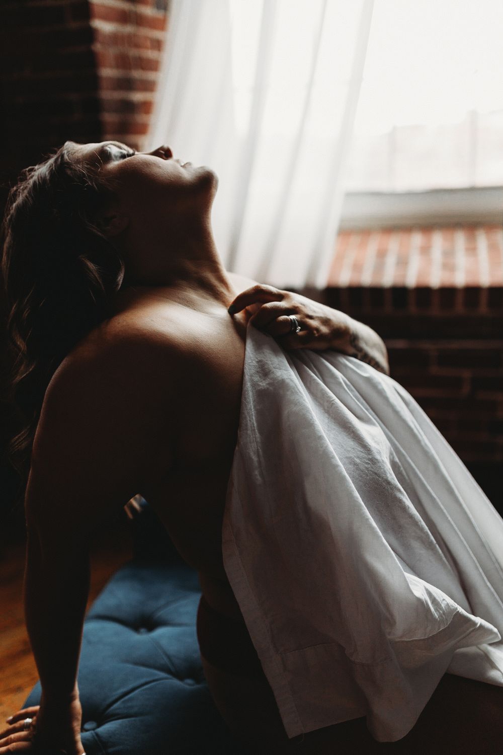 Dramatic boudoir photo showing person in white fabric leaning back against brick wall in moody lighting.