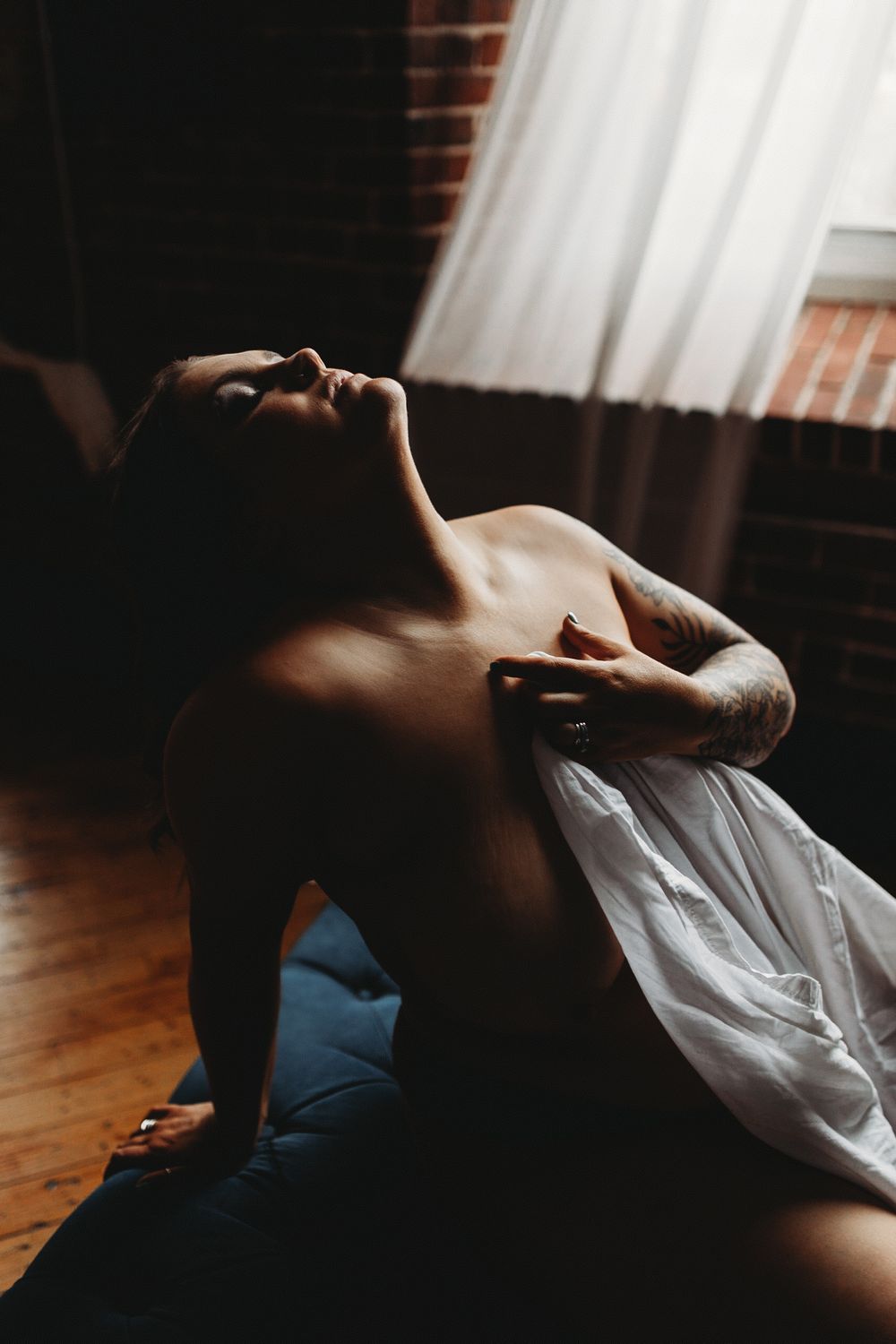 Dramatic moody boudoir photo with soft window light illuminating bare shoulders against brick wall and white curtains.