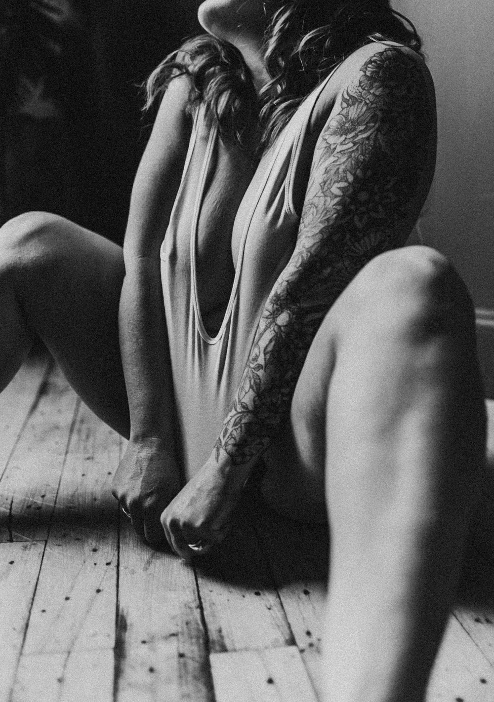 Black and white artistic shot of person wearing delicate clothing sitting on wooden floor with intricate arm tattoo.