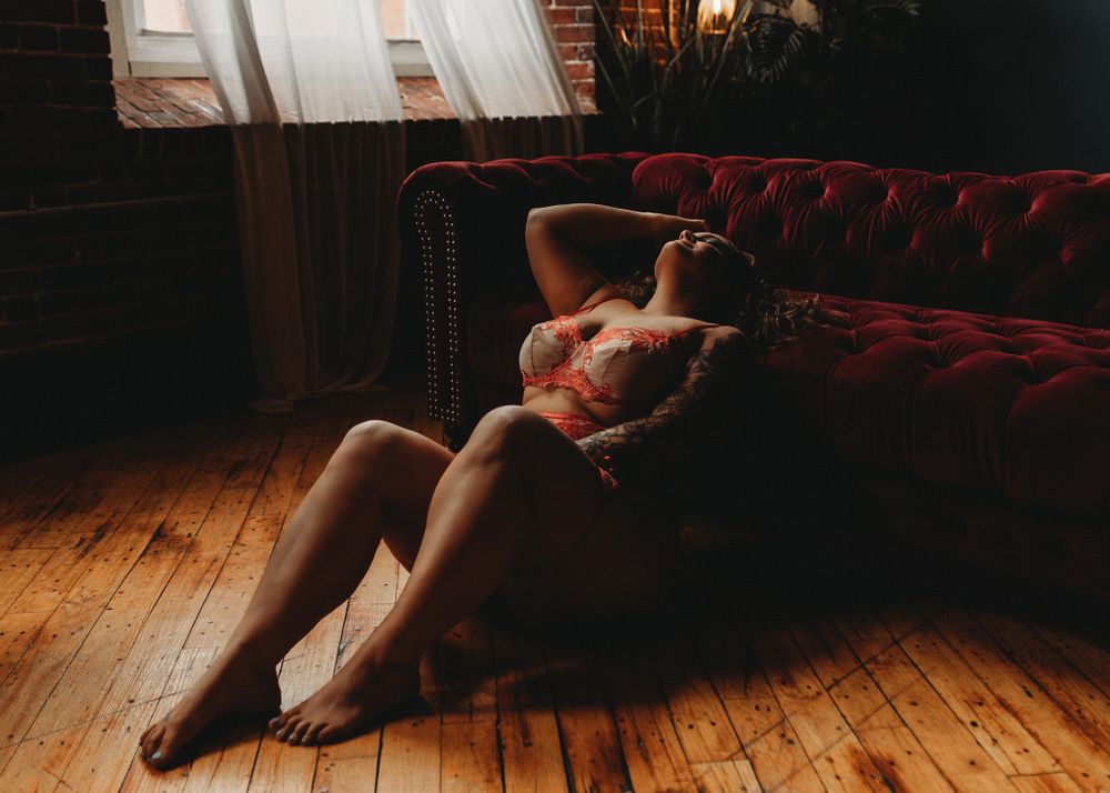 Person in lingerie reclining on hardwood floor next to red velvet couch in moody boudoir setting.