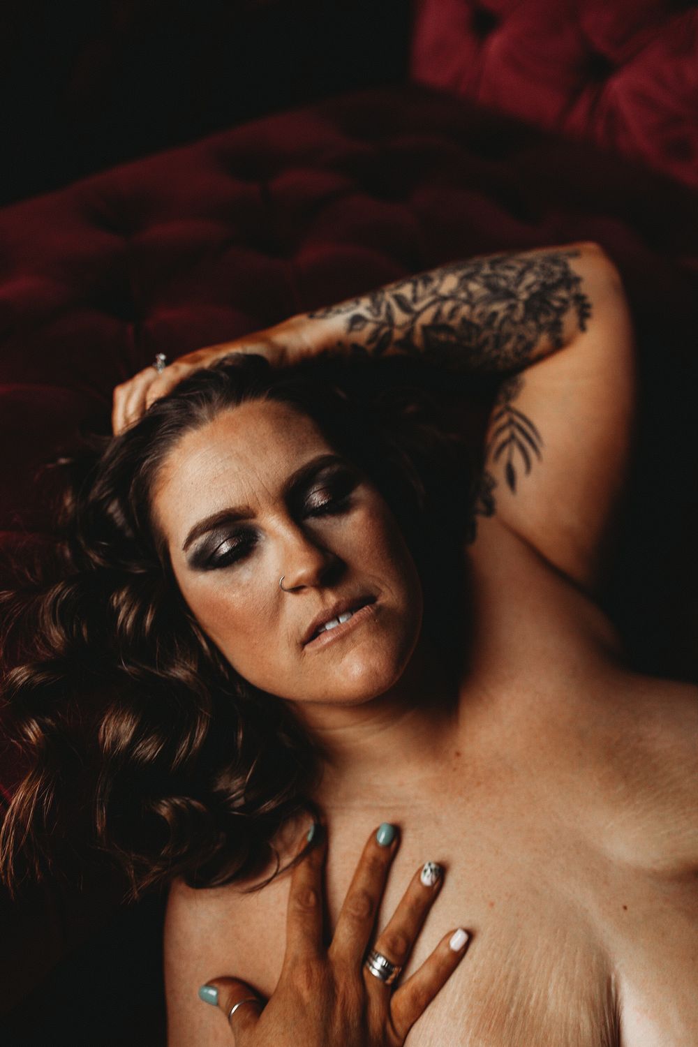 Intimate artistic portrait with moody lighting showcasing tattoos and jewelry against dark red background.