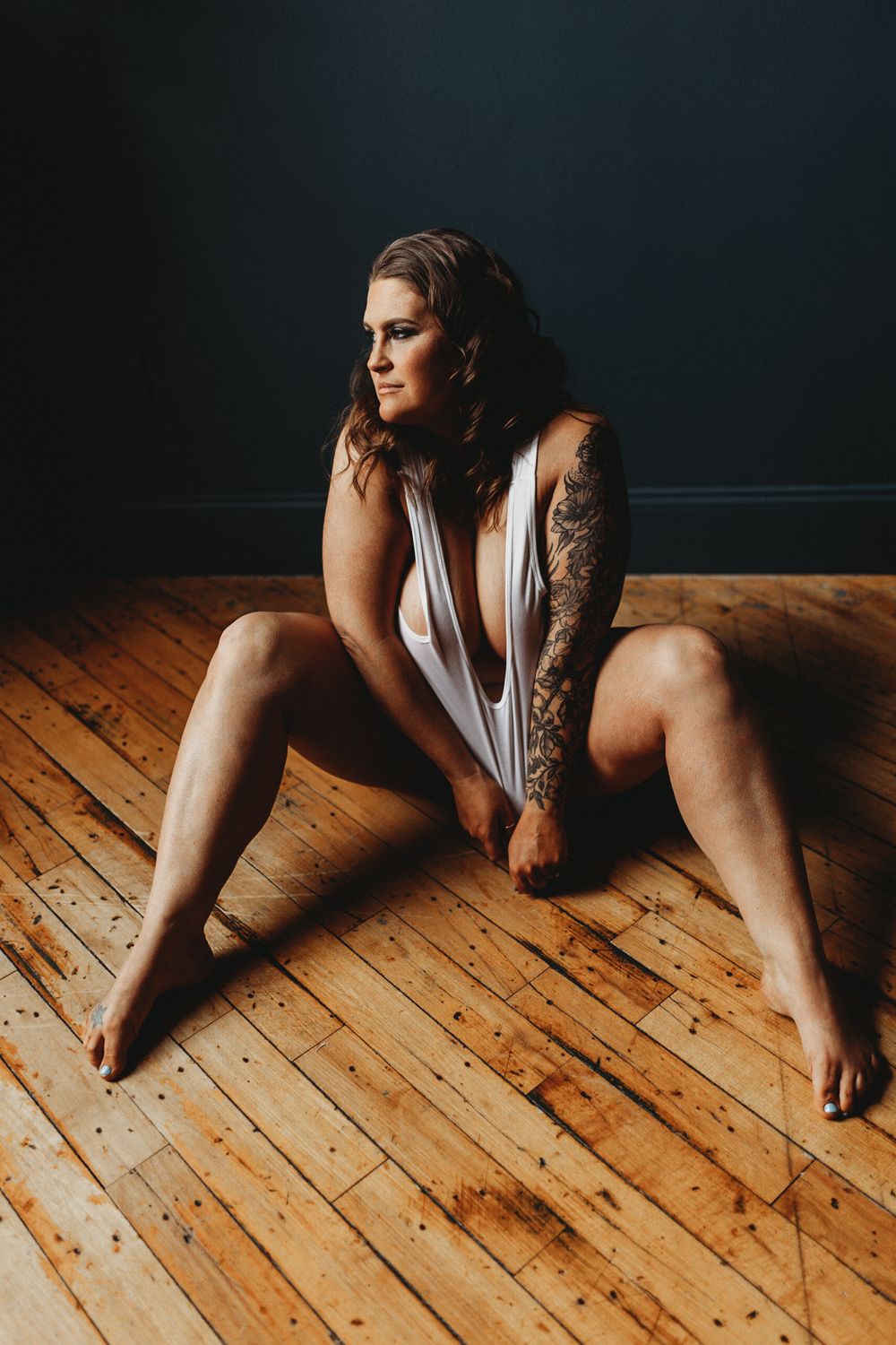 Dramatic portrait in white garment showing detailed arm tattoo against dark background on wooden floor.
