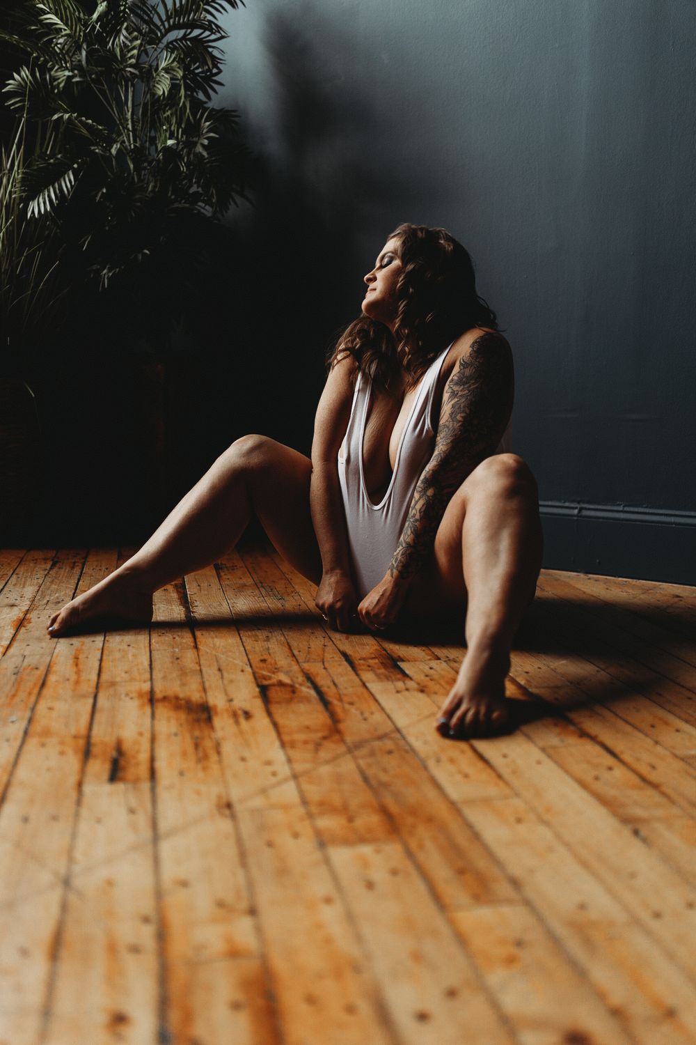 Artistic portrait on hardwood floor with dramatic natural lighting and plants.