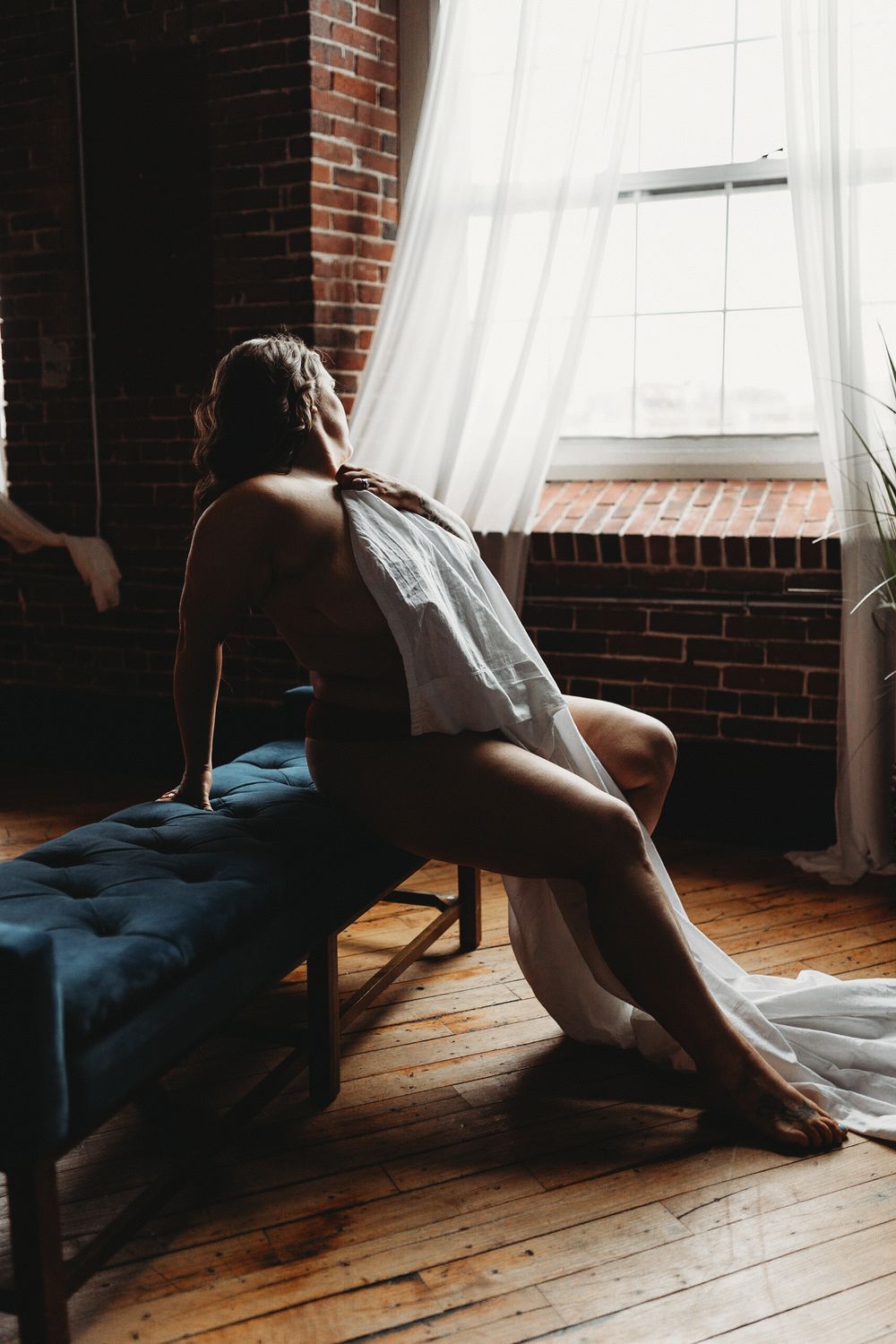Figure sits by large window with flowing white curtains in brick-walled room with hardwood floors and natural lighting.