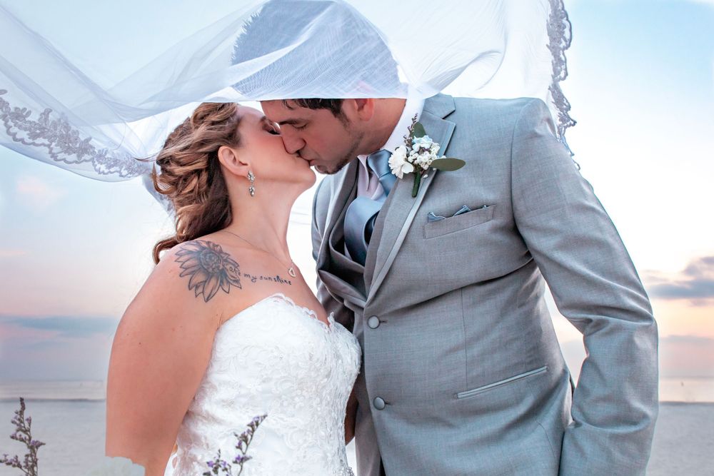 Wedding couple embraces on a beach at sunset with bridesmaids and groomsmen in gray and blue attire.