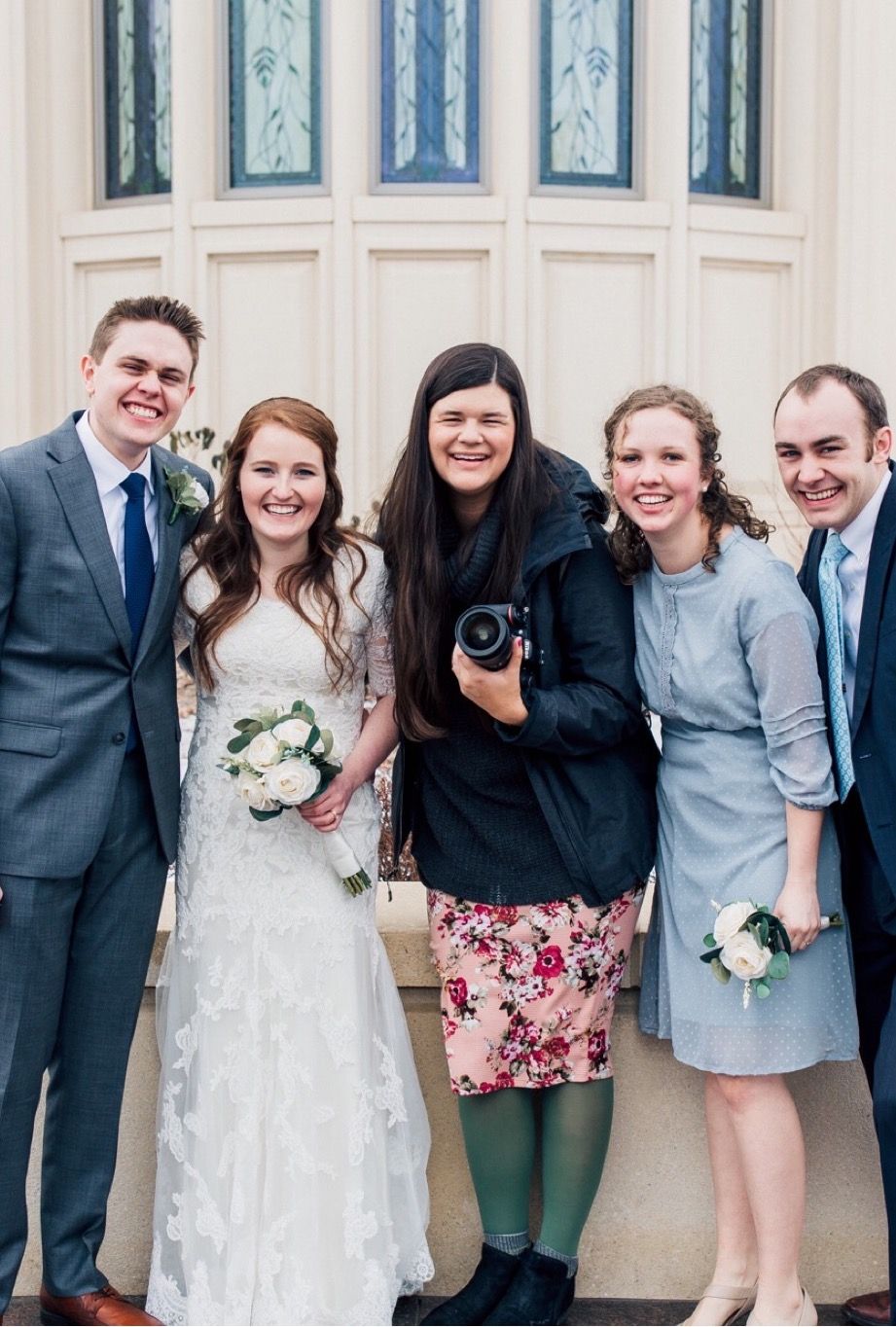 Professional photographer in floral skirt poses with wedding party in front of white building.