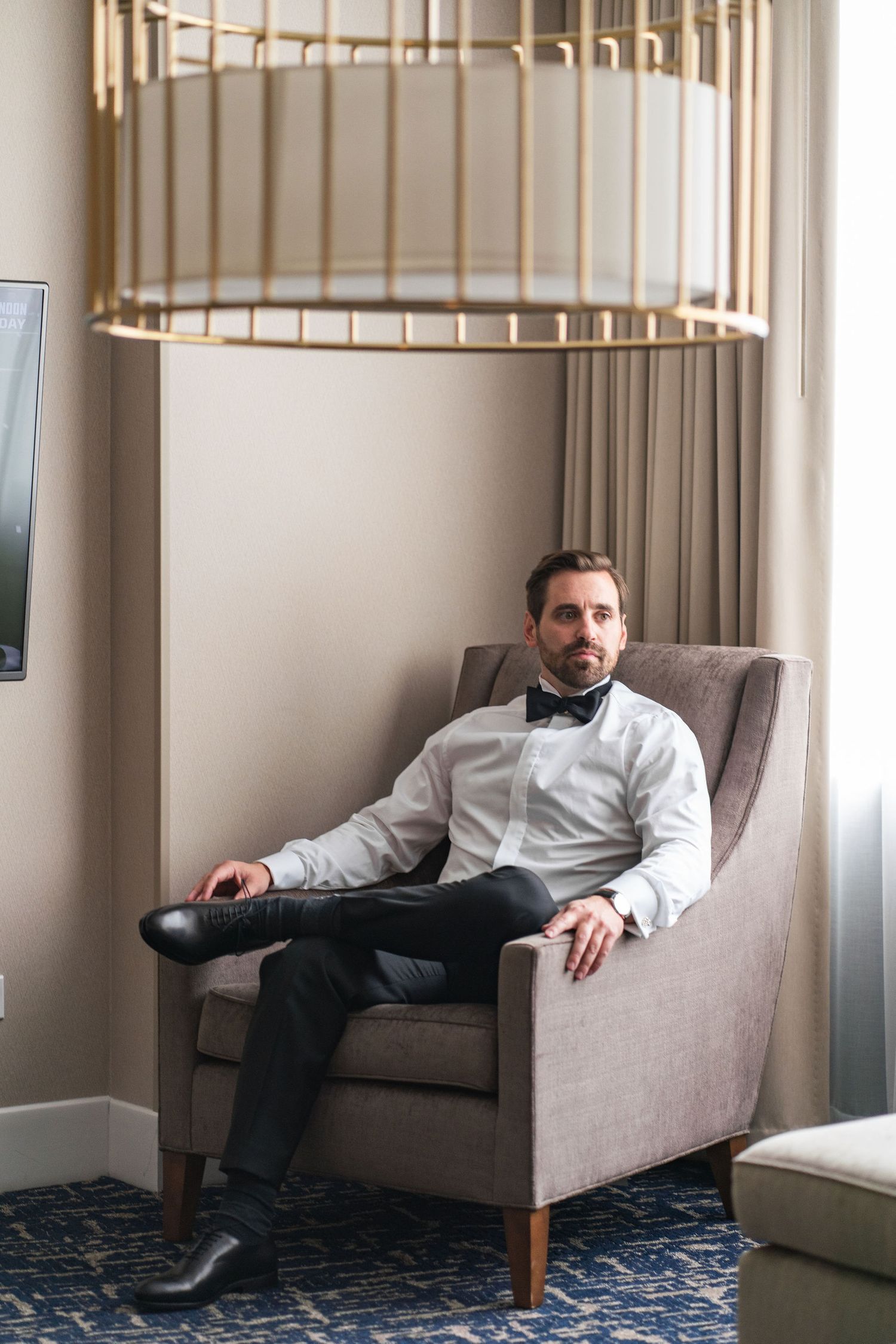 A well-dressed person relaxes in a gray armchair beneath a modern gold ceiling light fixture.