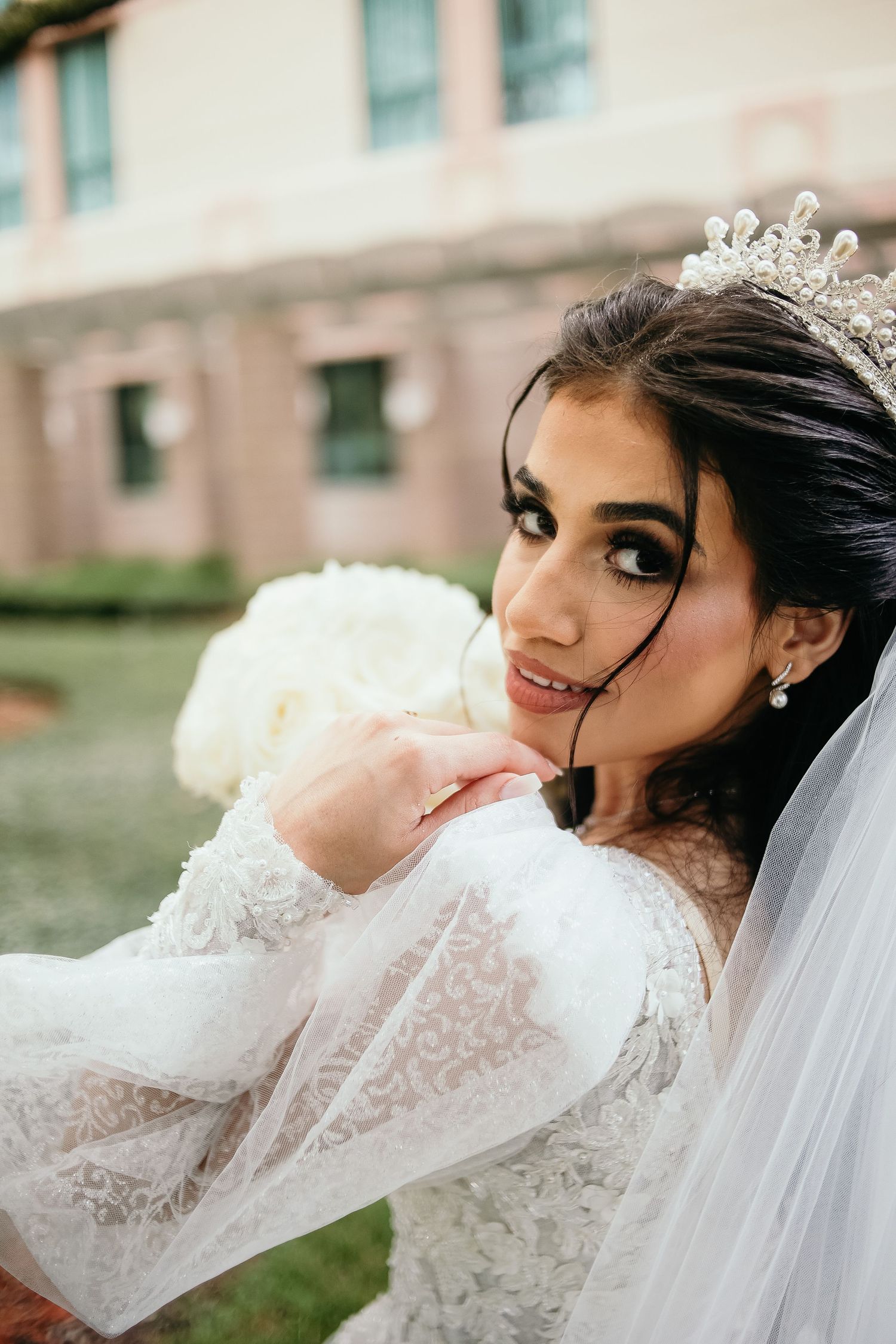 Elegant bride in white lace gown and veil poses gracefully outdoors.