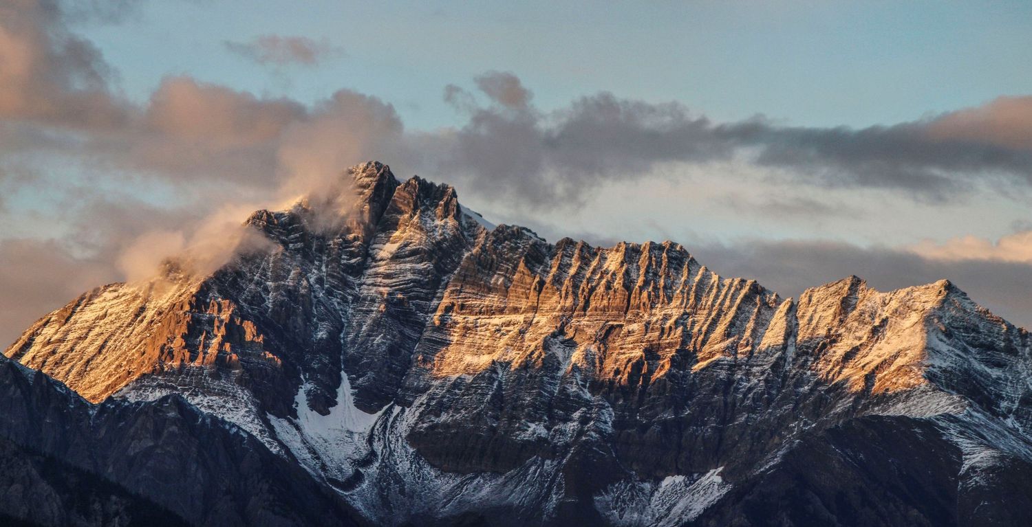 Snow-capped mountain peak bathed in golden sunset light with dramatic clouds above.
