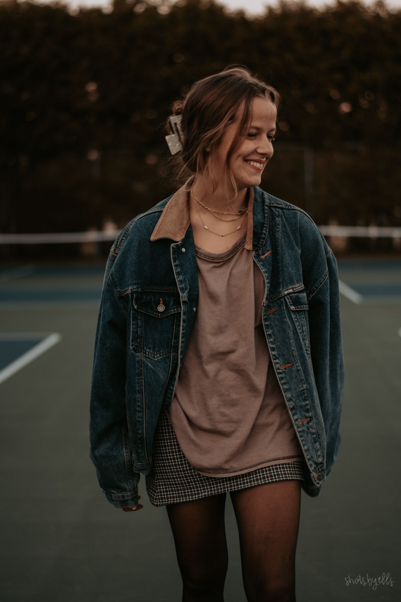 A casual autumn outfit featuring a denim jacket, brown top, and plaid skirt photographed at dusk on a tennis court.